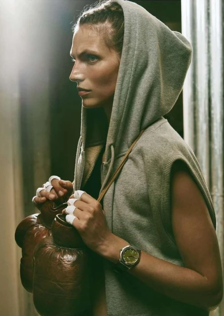 Woman with boxing gloves slung over shoulder, wearing a grey sleeveless hoodie and wristwatch; indoor, soft lighting, medium shot.