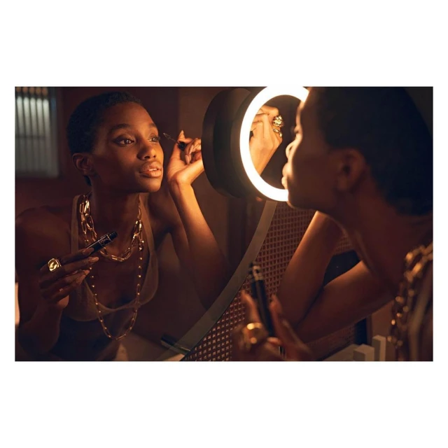 Woman applies makeup in front of a lighted mirror indoors. She wears a sleeveless top and gold jewellery around her neck and fingers.