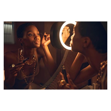 Woman applies makeup in front of a lighted mirror indoors. She wears a sleeveless top and gold jewellery around her neck and fingers.