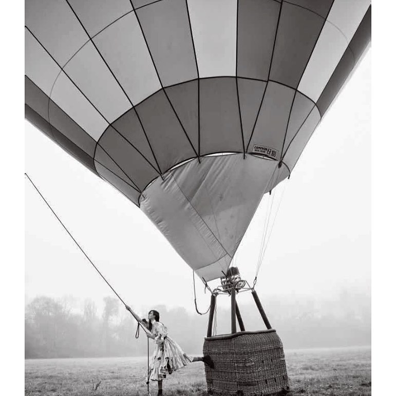Black and white photo of a woman in a floral dress standing next to a hot air balloon in a foggy field, captured from a low angle.