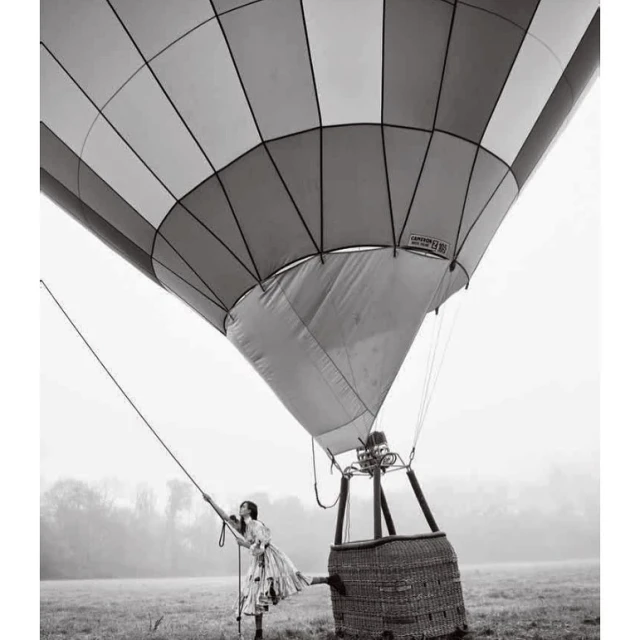Black and white photo of a woman in a floral dress standing next to a hot air balloon in a foggy field, captured from a low angle.
