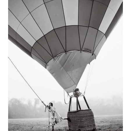 Black and white photo of a woman in a floral dress standing next to a hot air balloon in a foggy field, captured from a low angle.