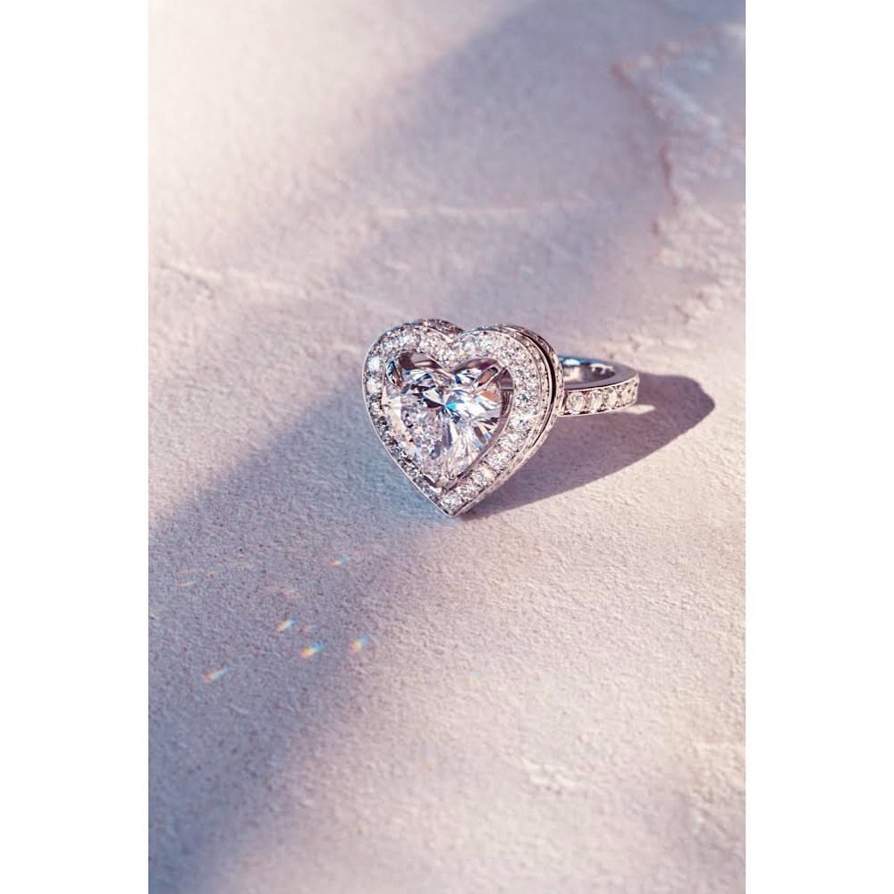 Close-up shot of a heart-shaped diamond ring with a halo setting, sparkling on a textured surface in soft, diffused lighting.