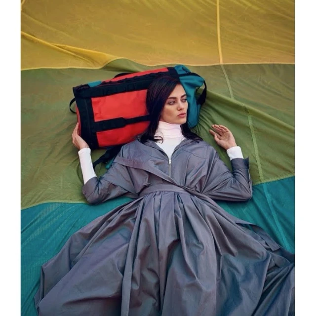 Woman in grey dress lies in tent, with backpack. Fashion editorial image with natural lighting and soft focus.