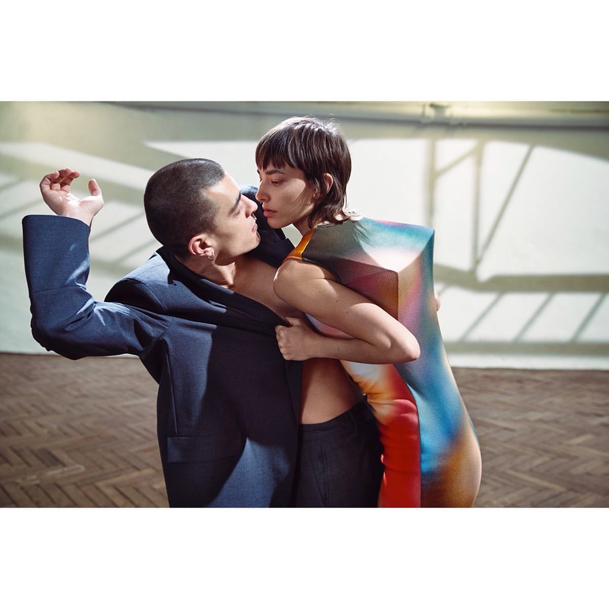 A fashion shot of a man and woman in an embrace. The woman wears a colorful dress, and the man sports a dark blue jacket with open torso.