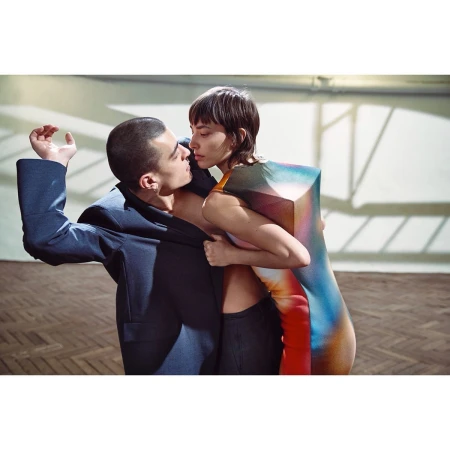 A fashion shot of a man and woman in an embrace. The woman wears a colorful dress, and the man sports a dark blue jacket with open torso.