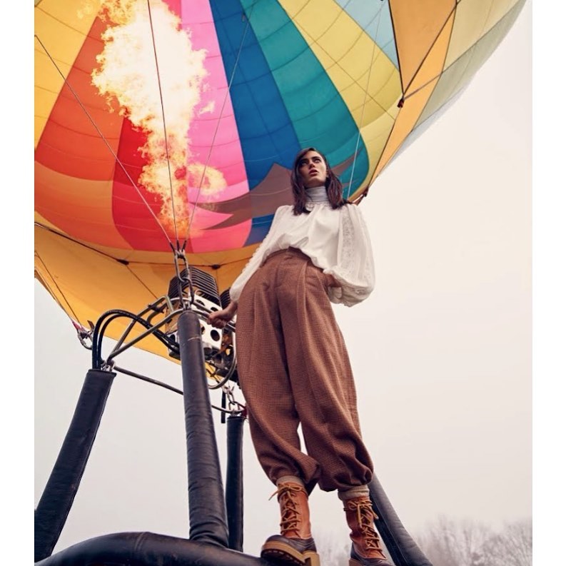 Woman standing in a hot air balloon basket with flames, wearing a white blouse, brown pants, and boots against a cloudy sky.