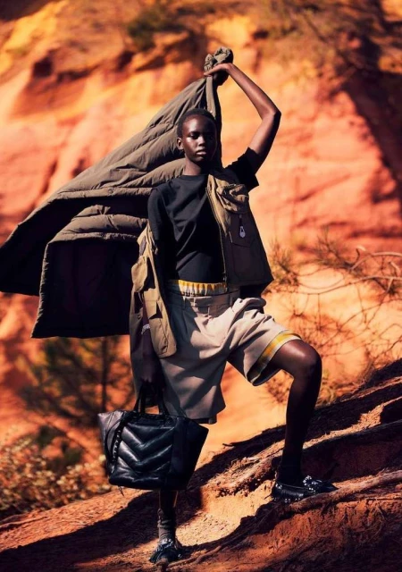 Fashionable Black woman outdoors in tan shorts, vest, carrying a black purse, holding a jacket, in front of an orange rock formation.