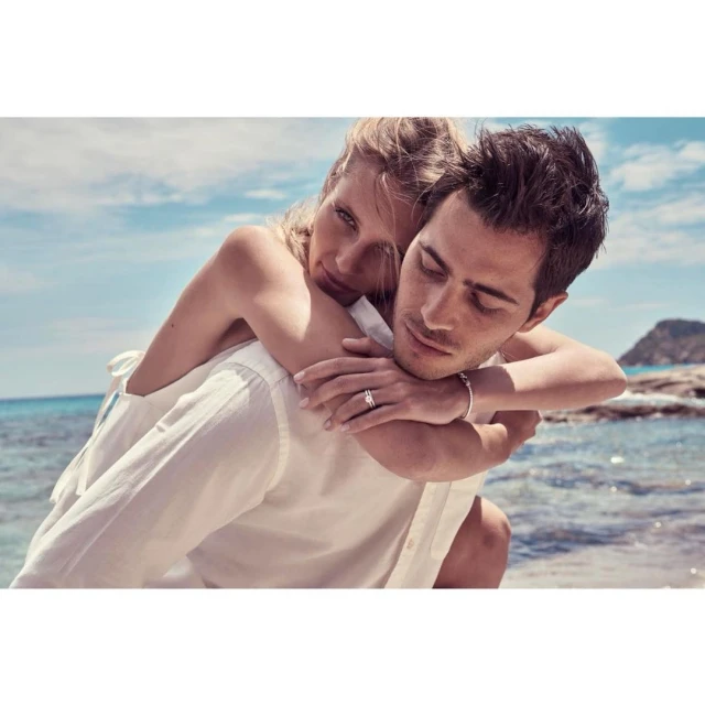 A woman rides piggyback on a man at the beach. The couple is wearing white clothing against an ocean background with rock formations.