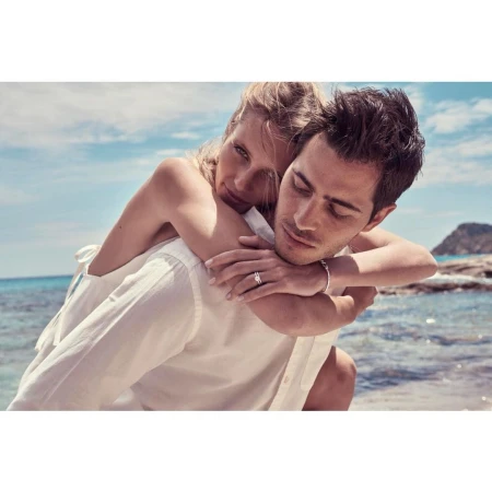 A woman rides piggyback on a man at the beach. The couple is wearing white clothing against an ocean background with rock formations.