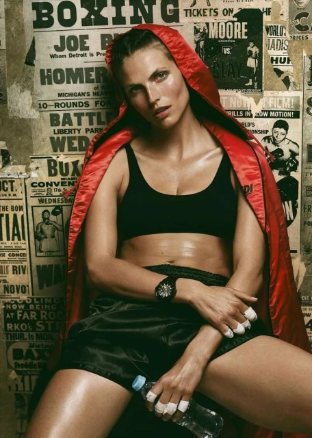 Female boxer in a red robe and black sports bra and shorts holding a water bottle, sitting in front of boxing posters.