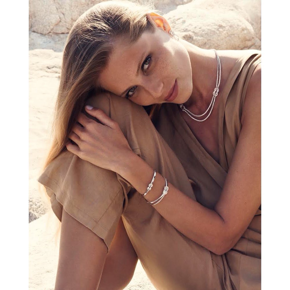 Adult woman wearing silver knot jewelry, including a bracelet and necklace, in an outdoor setting with neutral colors and soft light.