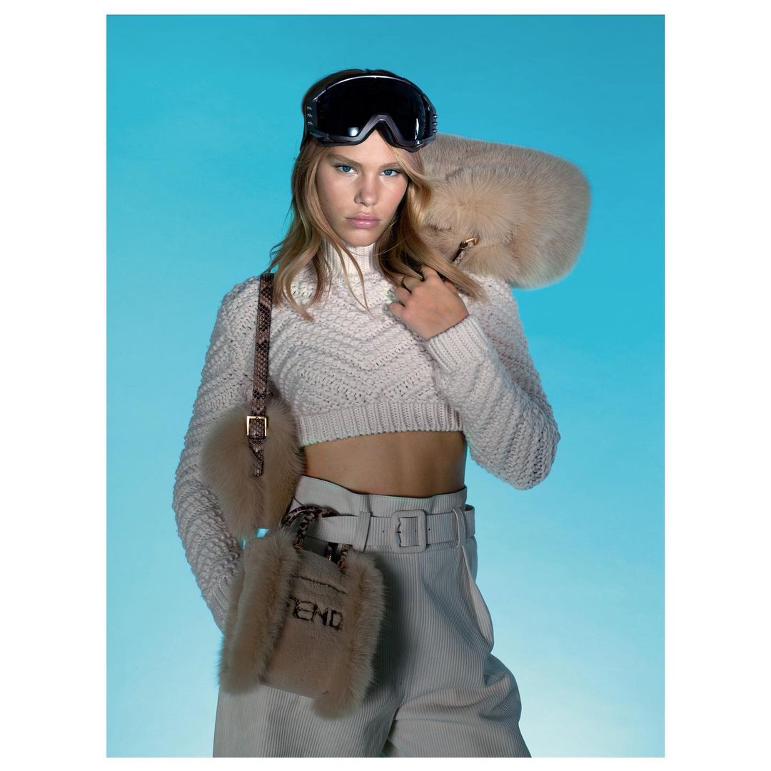 Fashion portrait of a young woman in ski goggles, cropped knit sweater, and pants, accented by fur accessories against a blue background.