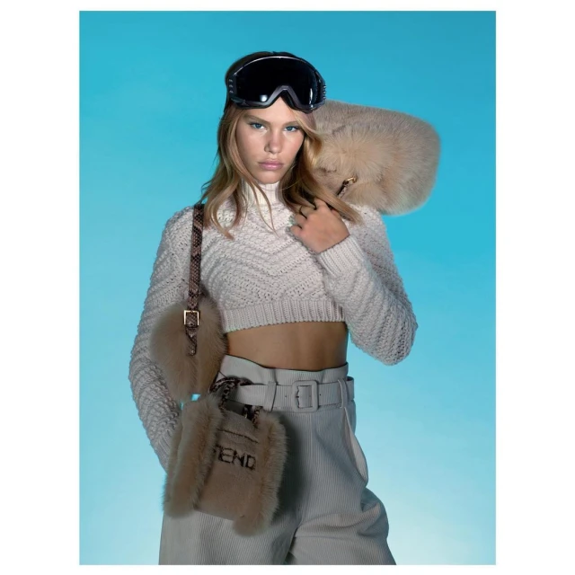 Fashion portrait of a young woman in ski goggles, cropped knit sweater, and pants, accented by fur accessories against a blue background.