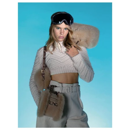 Fashion portrait of a young woman in ski goggles, cropped knit sweater, and pants, accented by fur accessories against a blue background.
