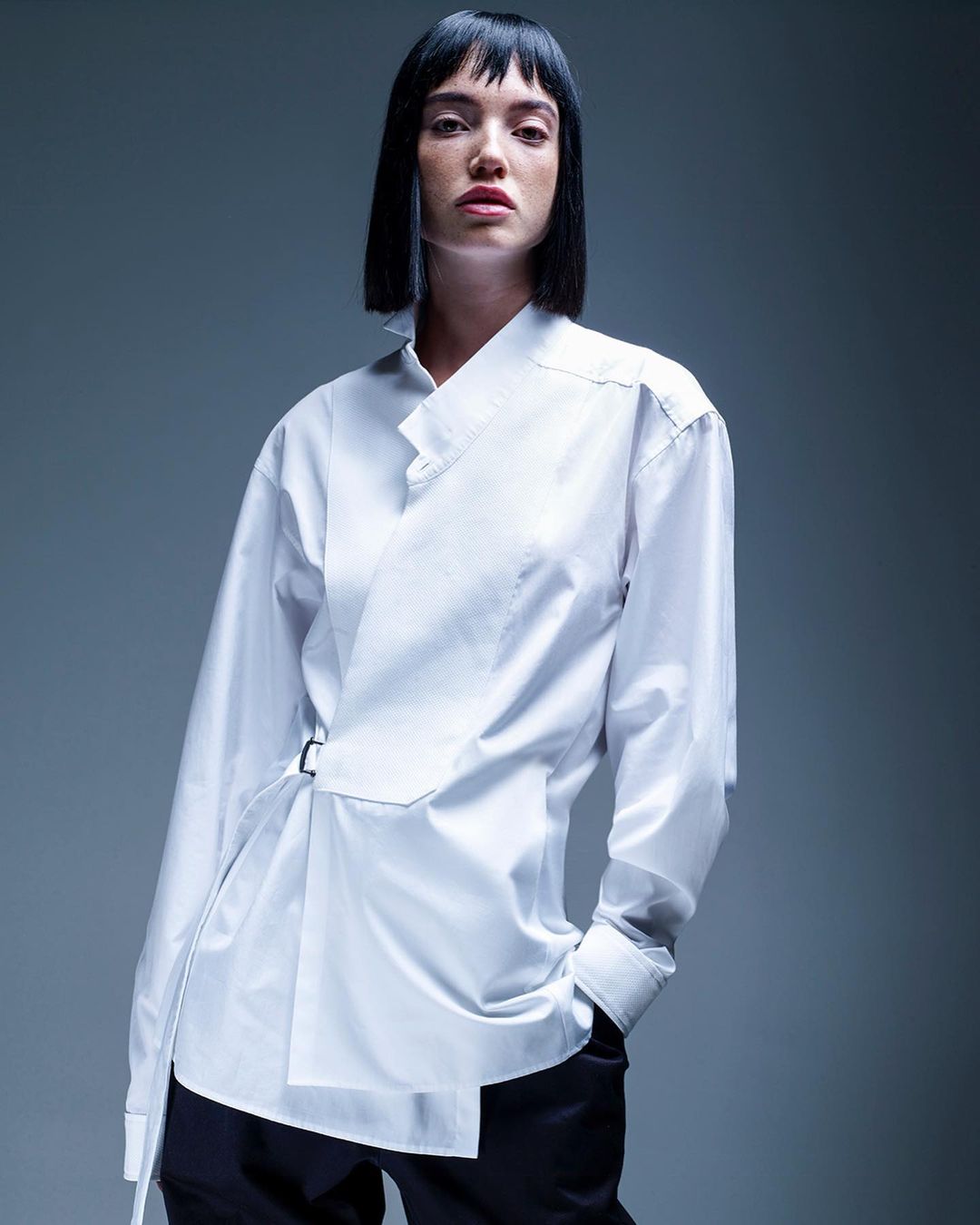 Studio shot of a young adult woman with dark hair wearing an asymmetric white shirt and dark pants against a gray background.