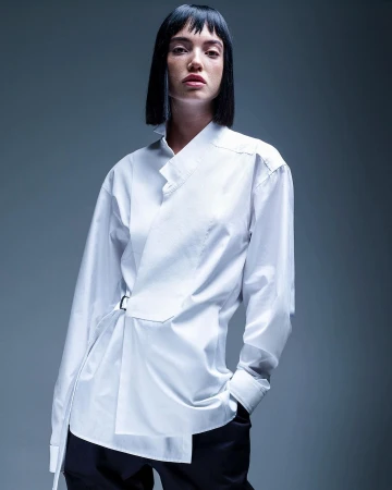Studio shot of a young adult woman with dark hair wearing an asymmetric white shirt and dark pants against a gray background.