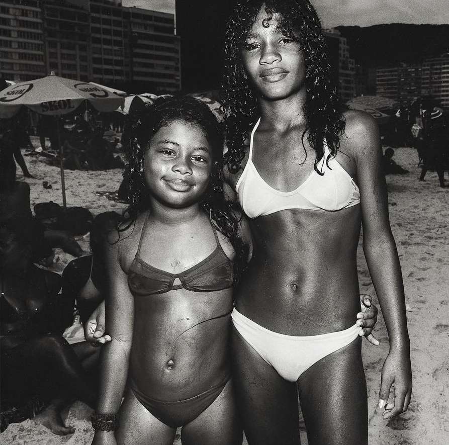 Two young girls in bikinis pose on a sandy beach with other people, buildings, and umbrellas in the background on a cloudy day.