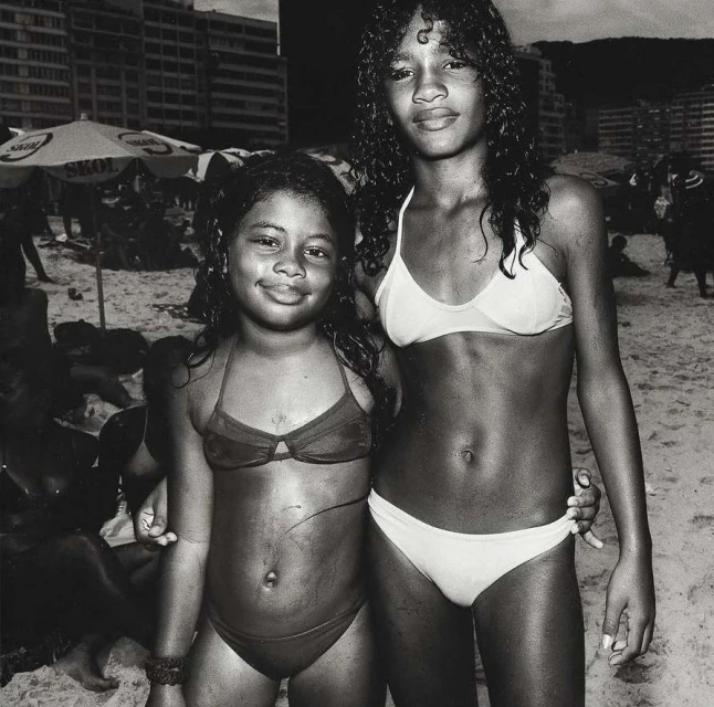 Two young girls in bikinis pose on a sandy beach with other people, buildings, and umbrellas in the background on a cloudy day.