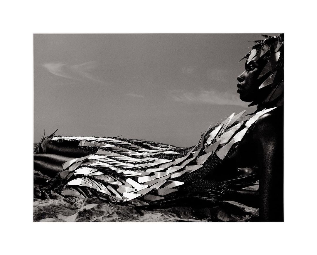Black and white portrait of a model wearing a reflective metallic shard headpiece and sculptural sequined dress outdoors against a sky.