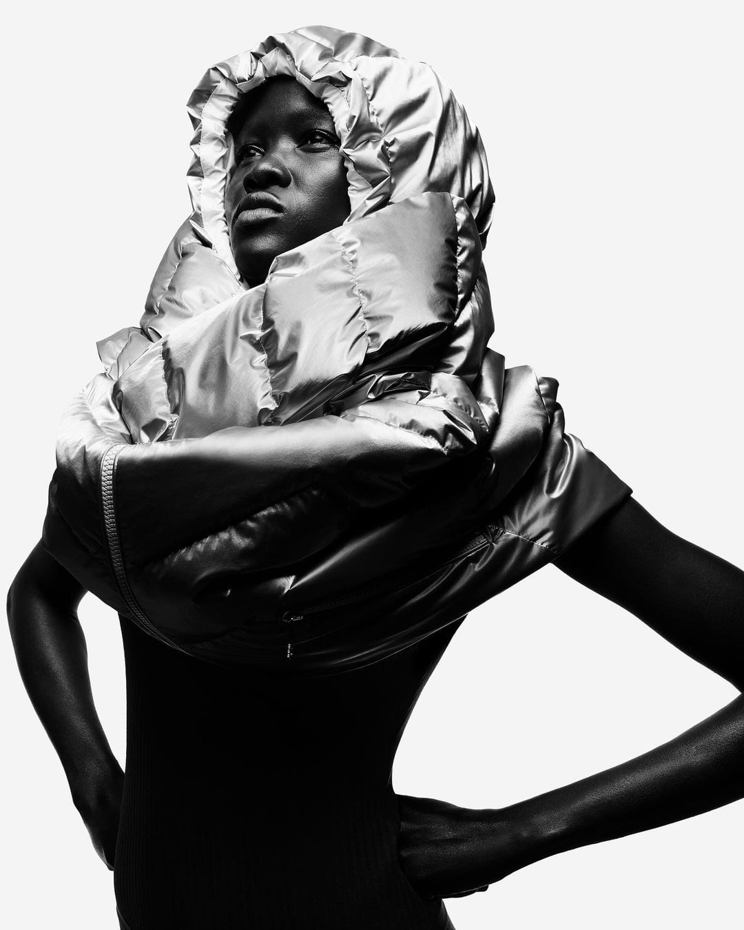 Striking fashion shot of an adult female in a black garment and shiny metallic puffer hood, hands on hips, against a white background.