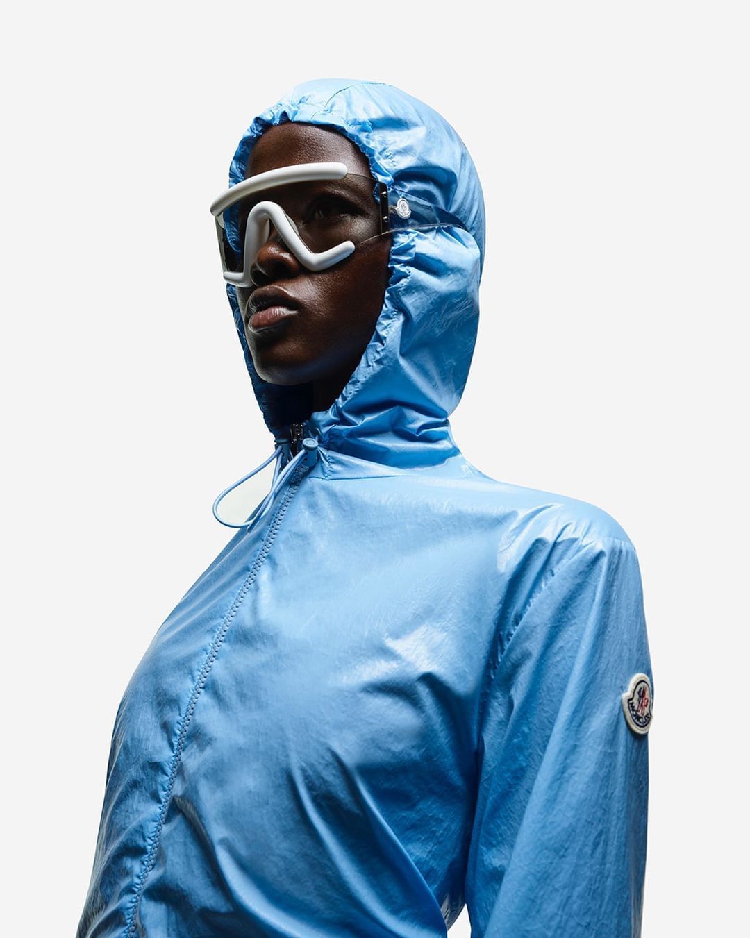 A woman models a shiny, light blue hooded jacket and white goggles against a white background, presented in a clean, fashion-forward style.