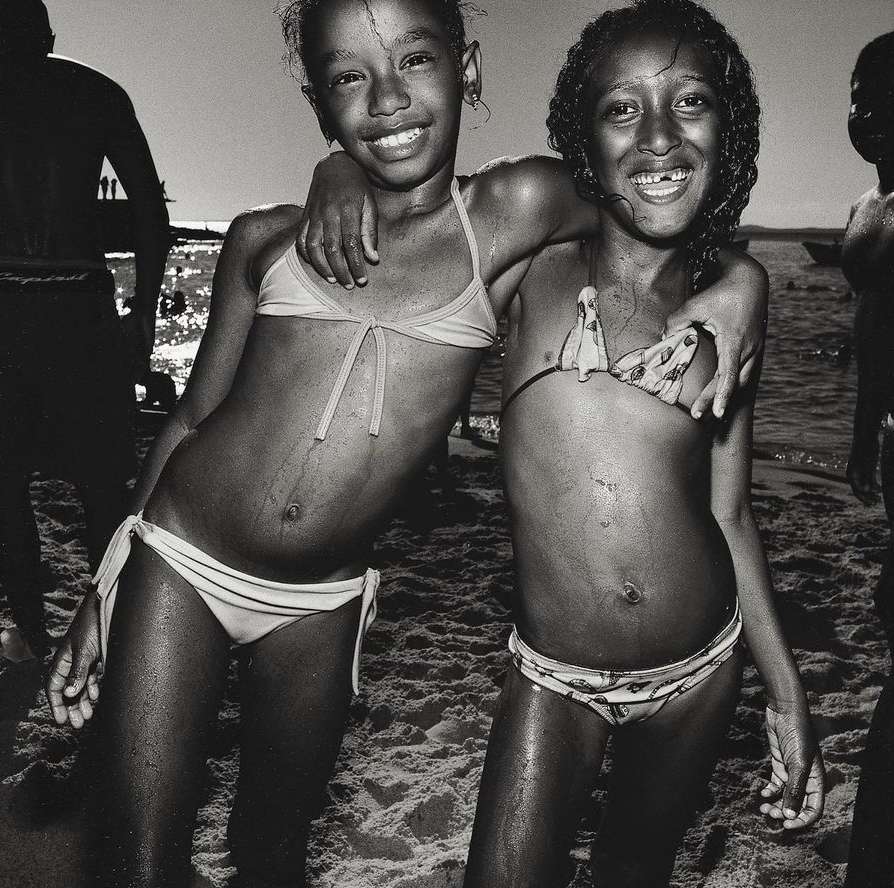 Two smiling girls in swimsuits embrace on a sandy beach in a black and white photograph, with other people and the sea visible in the background.
