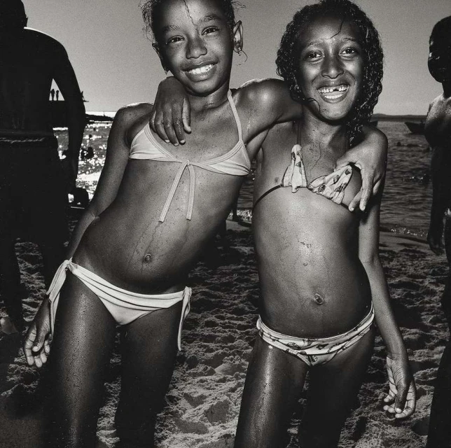 Two smiling girls in swimsuits embrace on a sandy beach in a black and white photograph, with other people and the sea visible in the background.