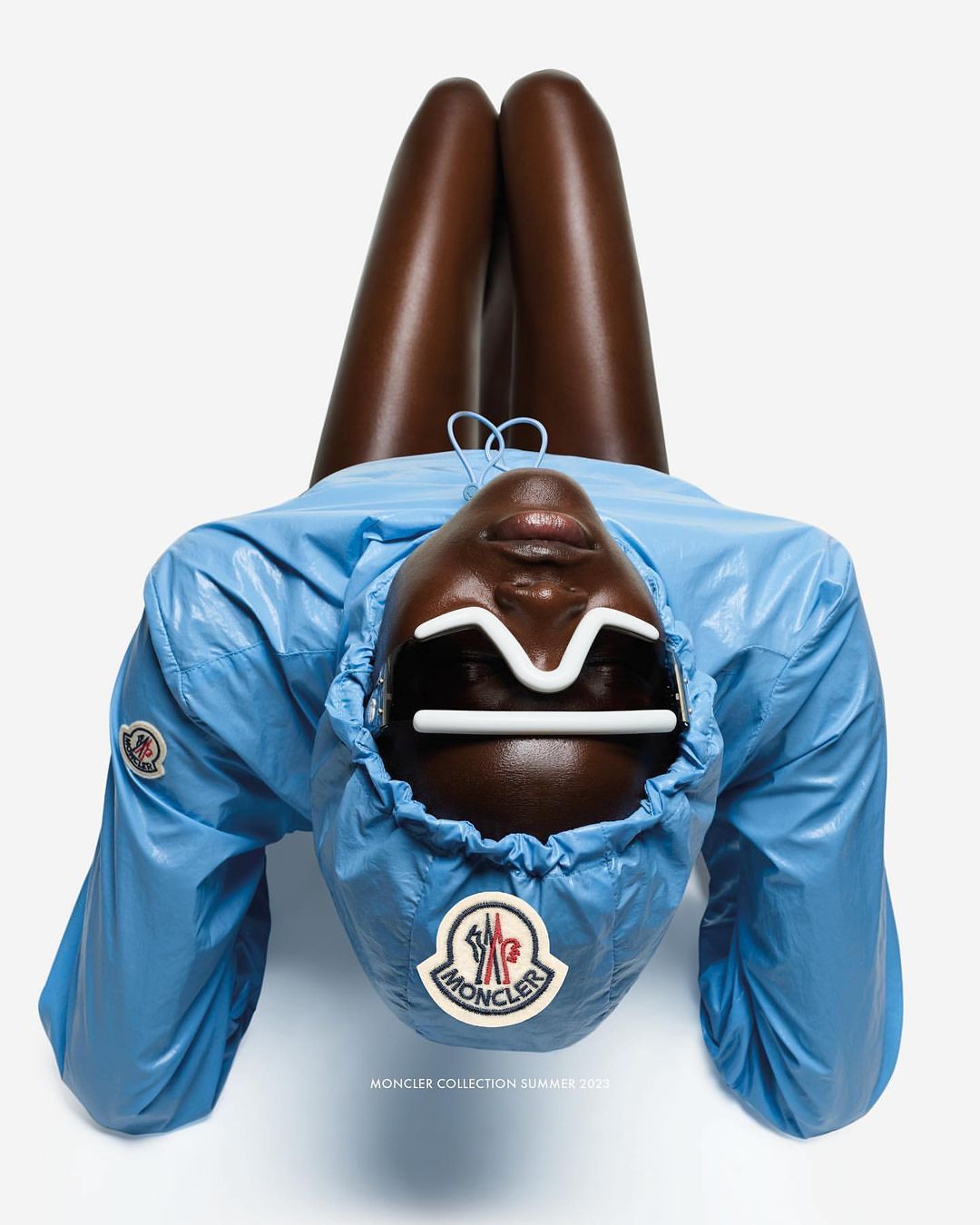 Fashion shot of a dark-skinned model wearing a blue jacket and unique glasses, lying with their legs up against a bright white background.