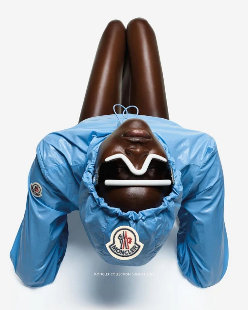 Fashion shot of a dark-skinned model wearing a blue jacket and unique glasses, lying with their legs up against a bright white background.