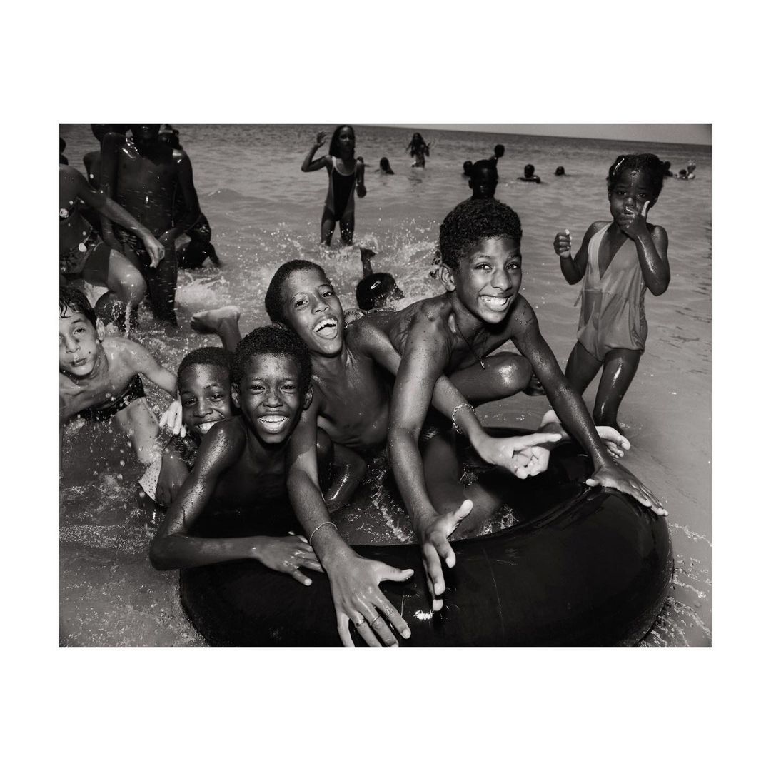 A group of Black children in water, some with an inner tube, smiling with a monochrome aesthetic creating a vintage, joyful beach scene.