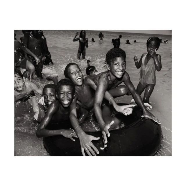 A group of Black children in water, some with an inner tube, smiling with a monochrome aesthetic creating a vintage, joyful beach scene.