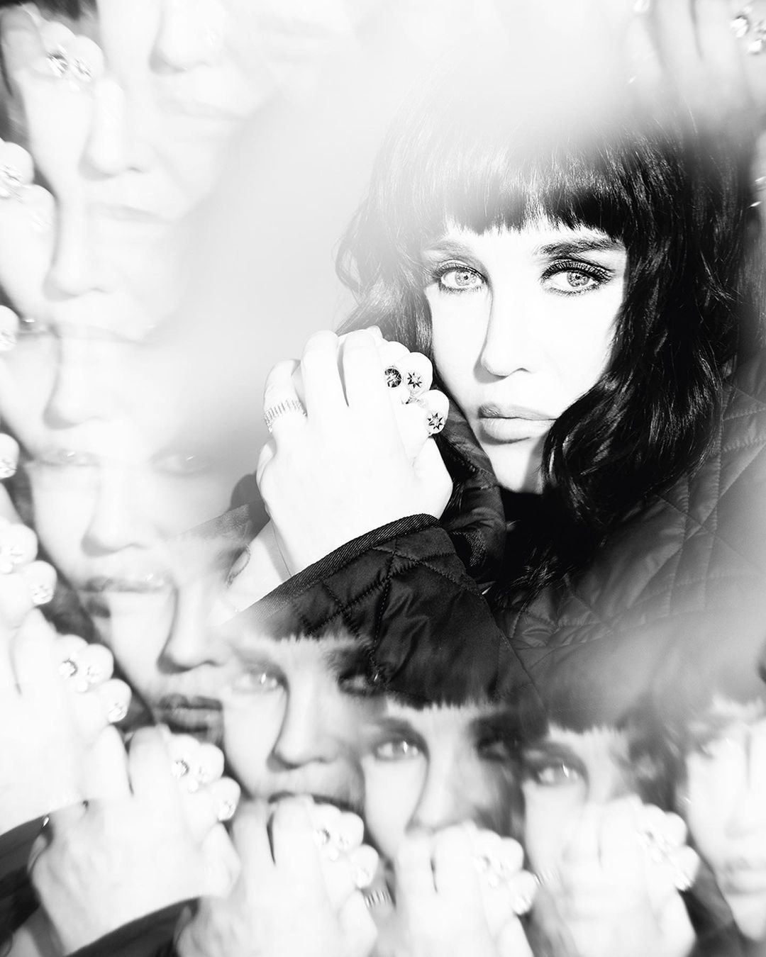 Black and white portrait of a woman with dark hair and bangs, wearing rings, with multiple superimposed face images and soft lighting.
