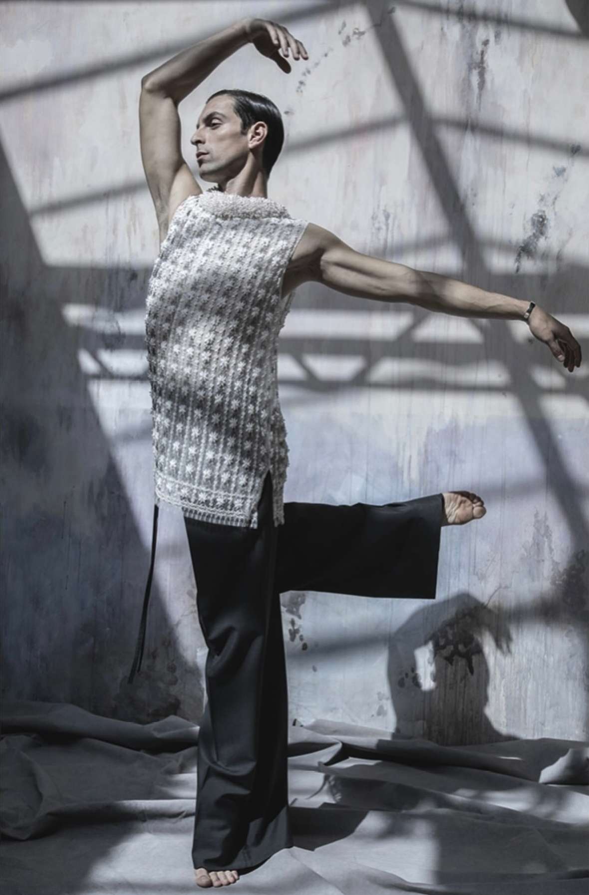 Man in textured sleeveless top and black pants in ballet pose, set against a textured wall and floor in a brightly lit studio.