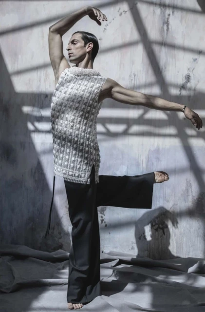 Man in textured sleeveless top and black pants in ballet pose, set against a textured wall and floor in a brightly lit studio.