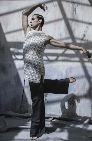 Man in textured sleeveless top and black pants in ballet pose, set against a textured wall and floor in a brightly lit studio.