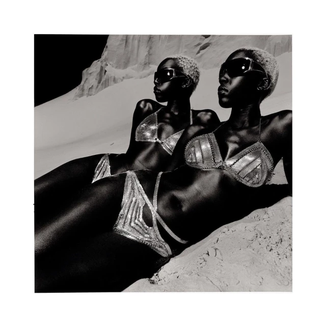 Two black female fashion models pose in shimmering bikinis on a bright, sandy beach. The black and white shot highlights the curves.