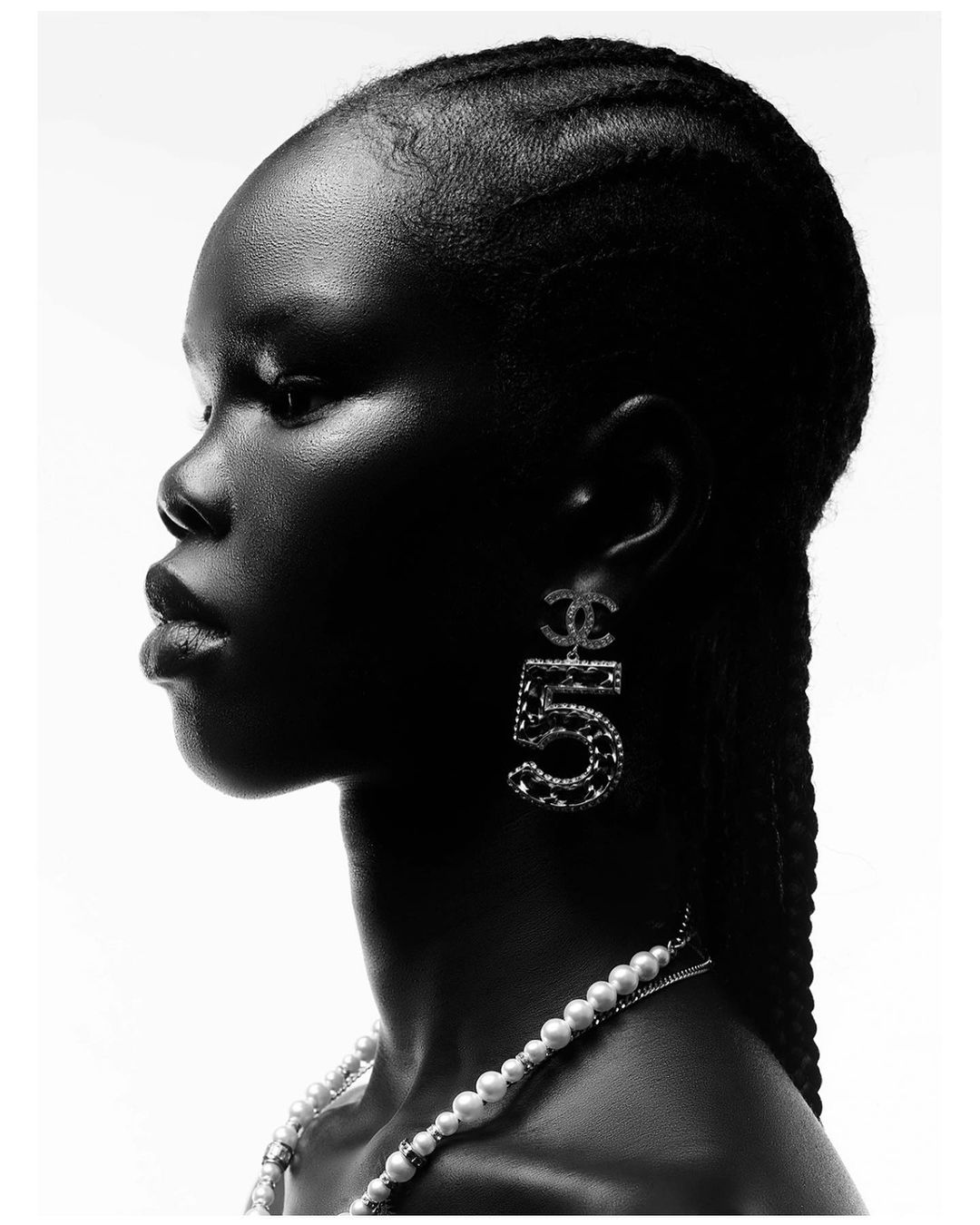 Striking monochrome portrait of a Black woman in profile, featuring braided hair, elegant pearl jewelry, and Chanel No. 5 earrings.
