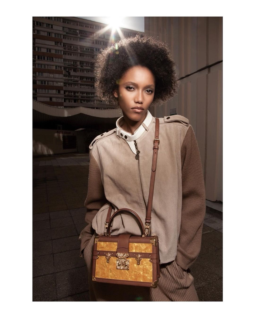 A woman with afro hair poses with a yellow and brown purse with gold hardware, wearing a matching tan jacket outdoors.