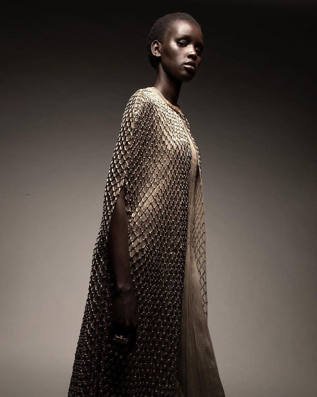 Elegant portrait of a model in a textured, beaded dress with cape. Dark background, studio lighting, and fashion-forward aesthetic.