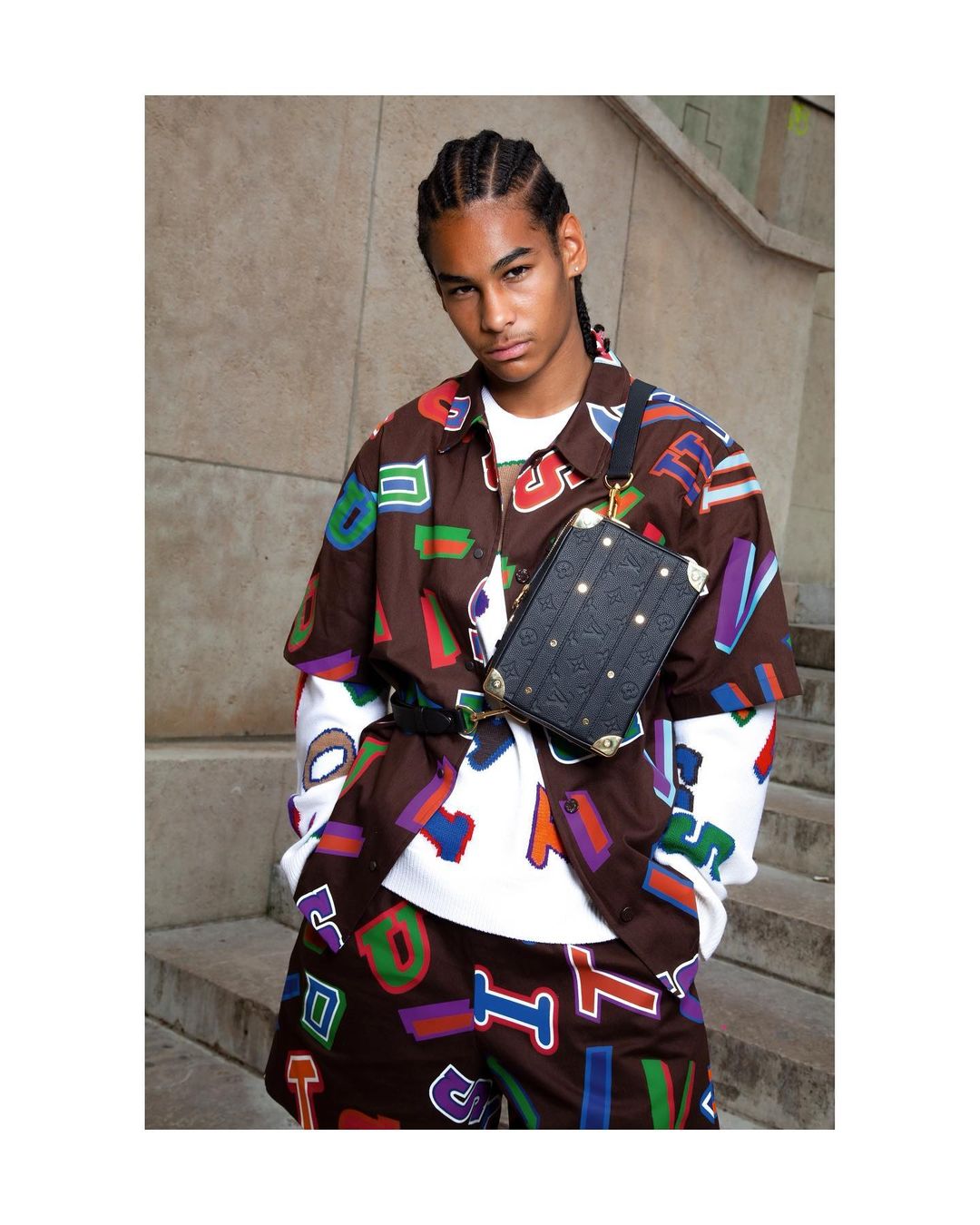 Male fashion model wears brown shorts and button-up shirt with multicolored letter graphics and black messenger bag in urban setting.
