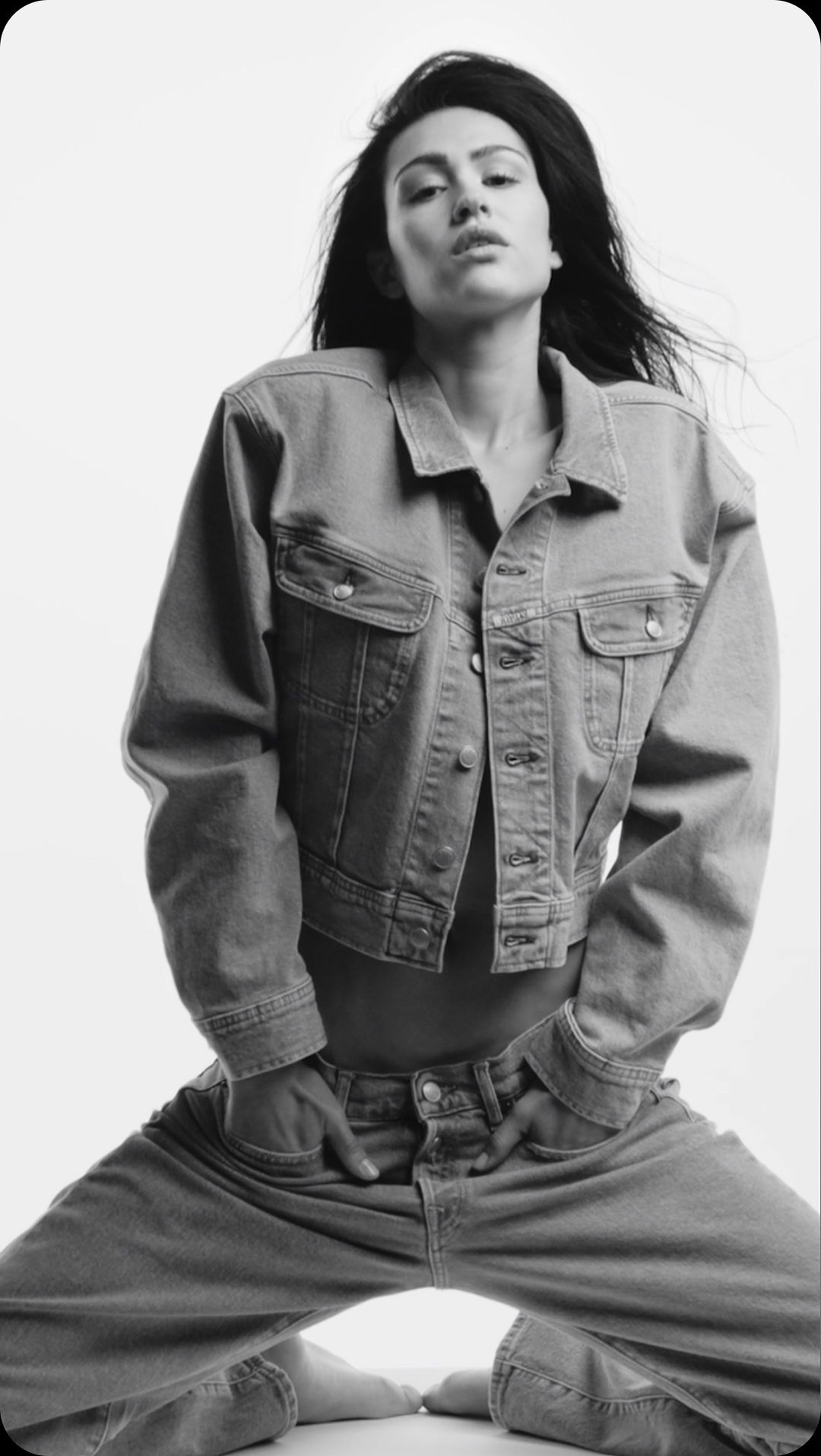 Monochrome portrait of a woman kneeling, wearing a denim jacket and jeans, against a bright white backdrop in a photo studio.