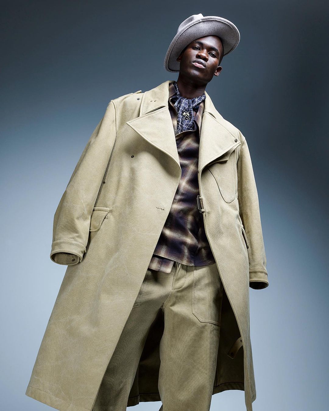 An adult male model poses wearing a light-khaki hat, tie-dye collared shirt, matching pants, and khaki trench coat in a studio setting.