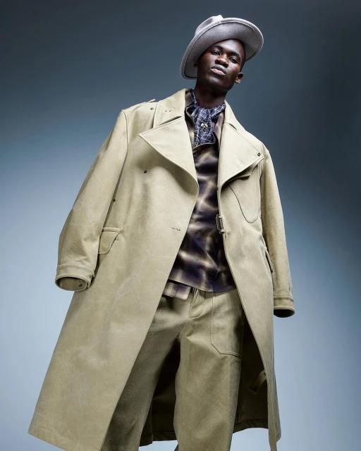 An adult male model poses wearing a light-khaki hat, tie-dye collared shirt, matching pants, and khaki trench coat in a studio setting.