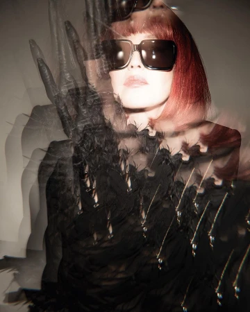 Portrait of a red-haired woman with sunglasses and a black, feathered top with water droplets. The scene is a light-toned indoor studio.
