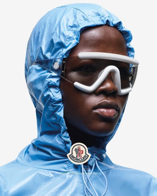 Close-up of a woman with dark skin wearing a shiny blue hooded jacket and white futuristic glasses against a white background.