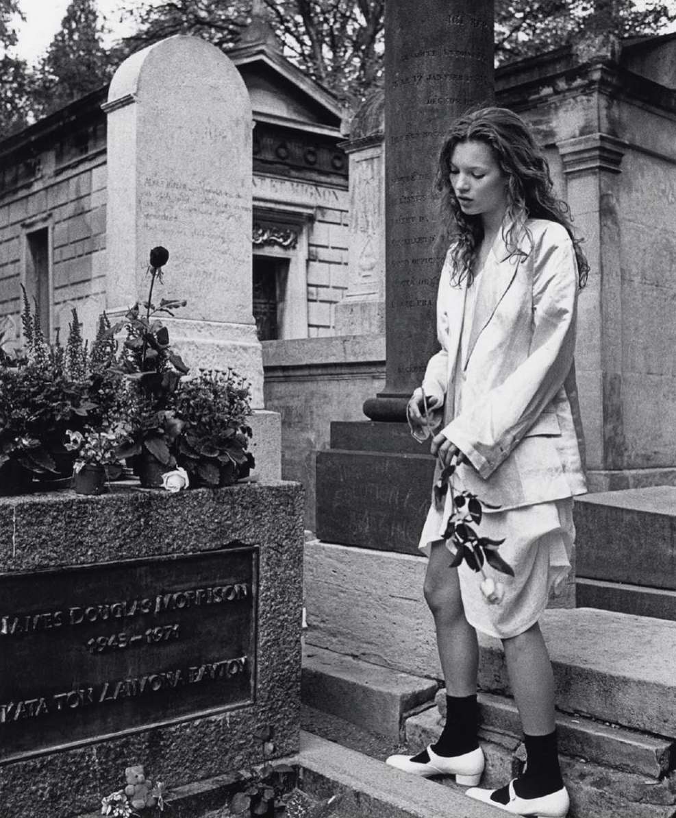 Black and white photograph of Kate Moss standing next to a tombstone in a cemetery holding a single black rose. 