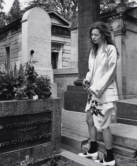 Black and white photograph of Kate Moss standing next to a tombstone in a cemetery holding a single black rose. 