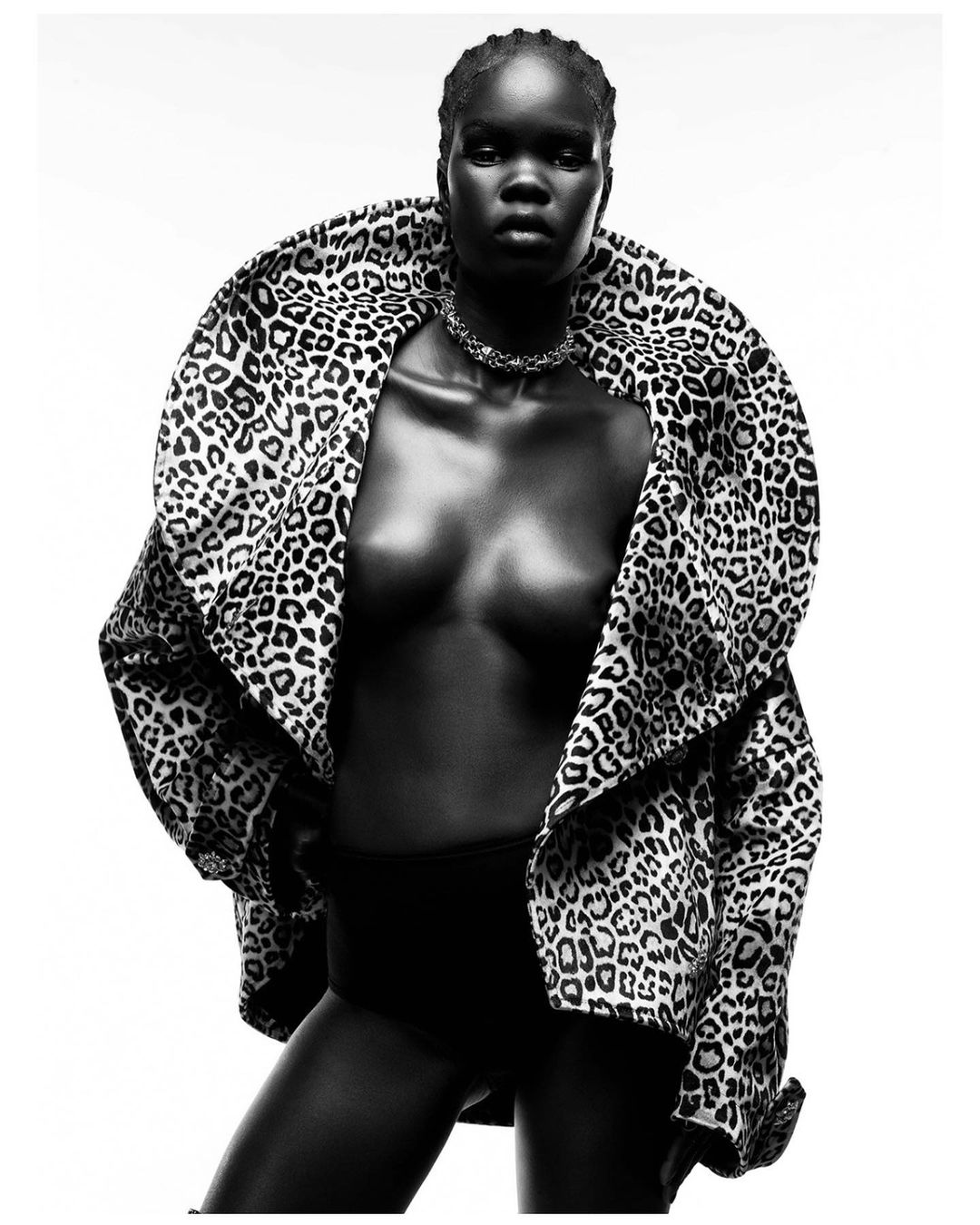 Striking black and white portrait of a Black woman wearing a leopard-print jacket and necklace against a bright white backdrop in a studio setting.