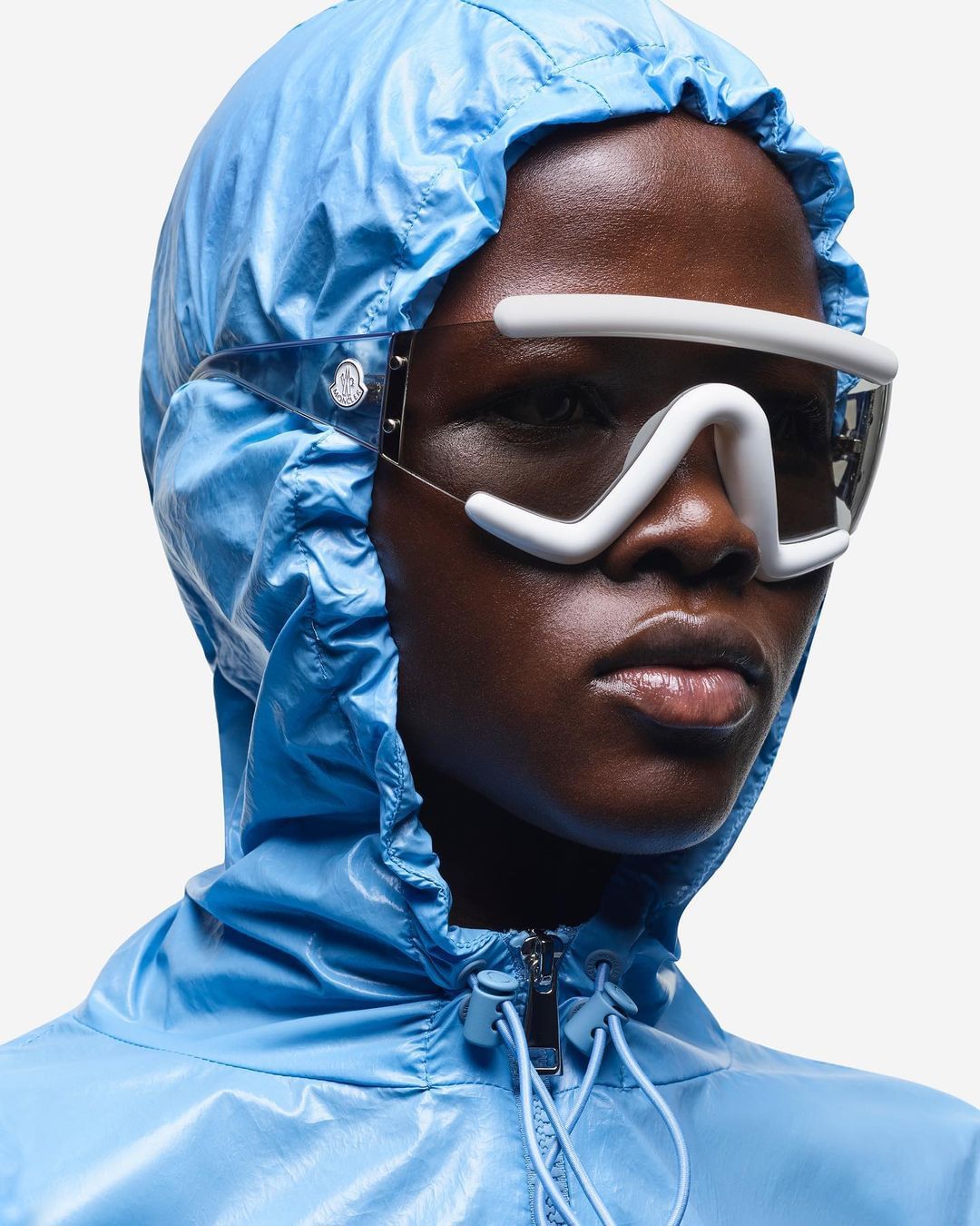A model with a dark complexion wearing a light blue hooded jacket and distinctive white eyewear against a white backdrop.